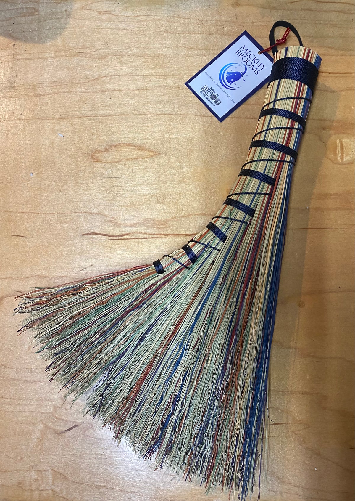 Broom with multicolored bristles on a wooden surface, featuring a label.
