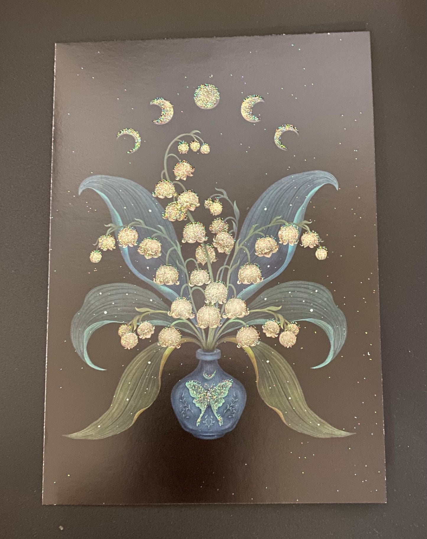 A square art print or card featuring a stylized bouquet of cream-colored flowers in a blue vase, with pale blue leaves and gold crescent moons against a dark brown speckled background.