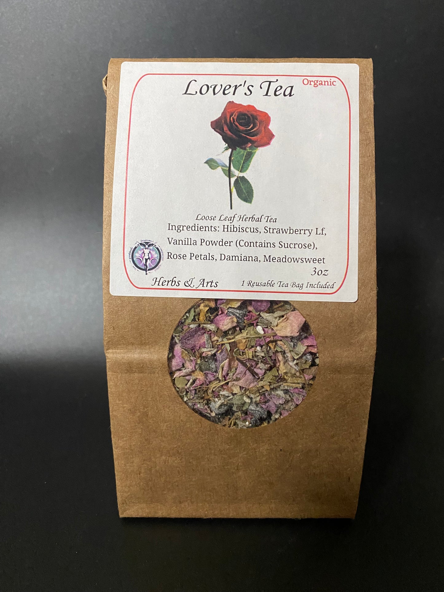 A kraft paper pouch of 'Lover's Tea' herbal tea with a white label featuring a red rose illustration and a circular cutout showing the dried flower blend inside, set against a dark background.