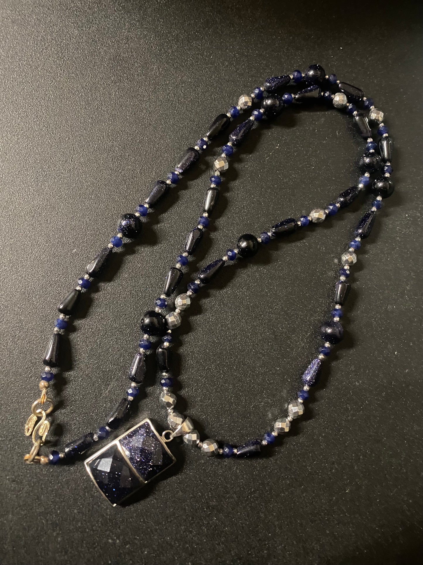A beaded necklace featuring dark blue goldstone, blue faceted beads, and silver-toned spacer beads, with a square dark blue pendant set in silver, displayed on a dark textured background.