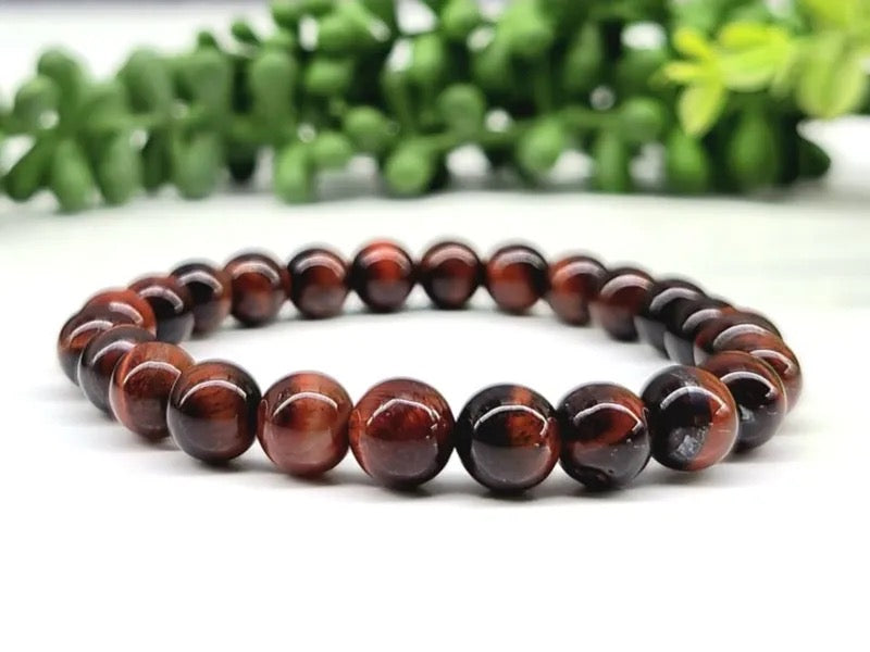 A beaded stretch bracelet made of polished red tiger's eye beads, displayed on a white surface with a blurred green plant in the background.