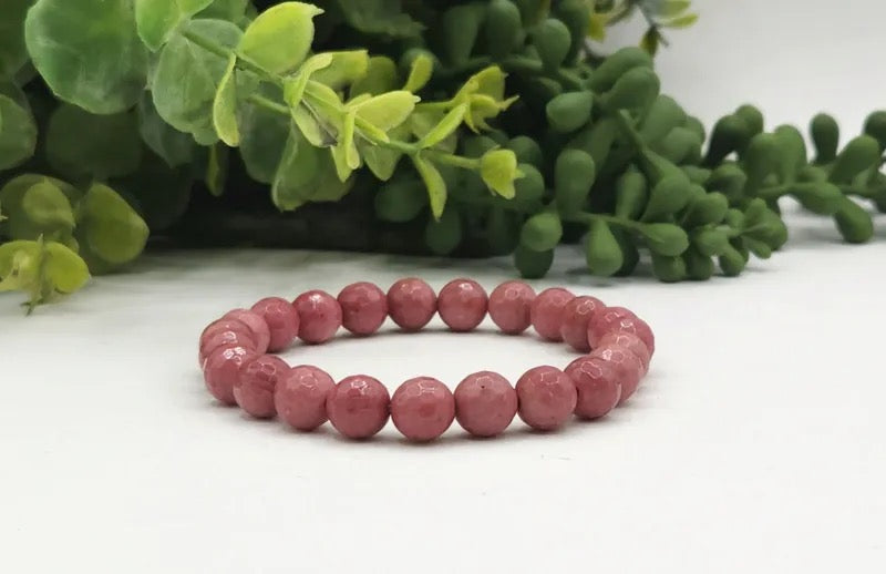 A beaded stretch bracelet made of faceted, round dusty rose beads, displayed on a white surface with green foliage in the background.