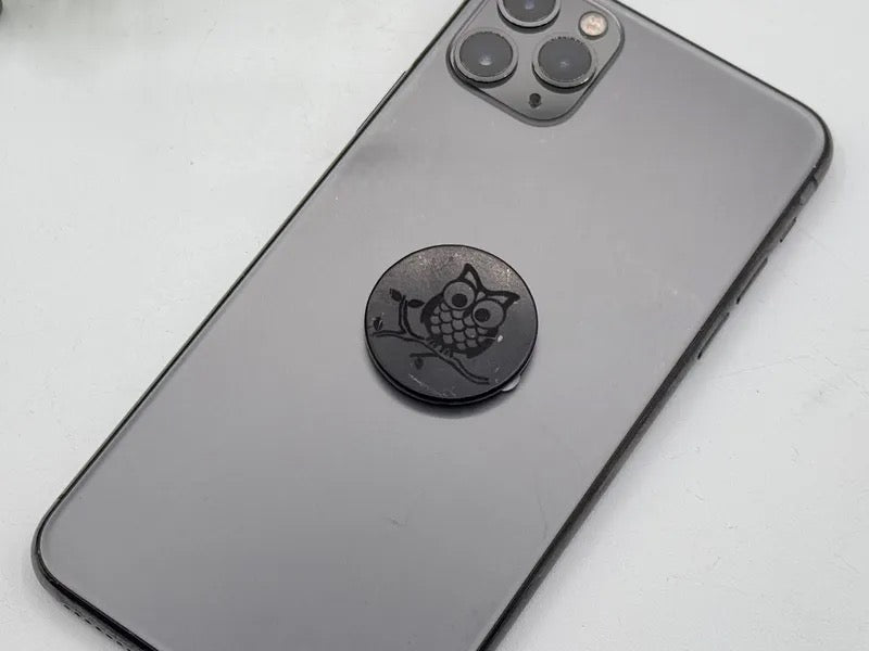 A black circular phone stand with an engraved owl design, shown attached to the back of a dark gray smartphone on a light gray surface.