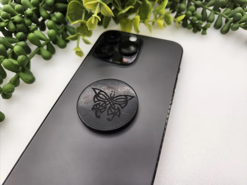 A black smartphone with a circular black tile featuring an engraved butterfly design, placed on a white surface with green plant leaves in the background.