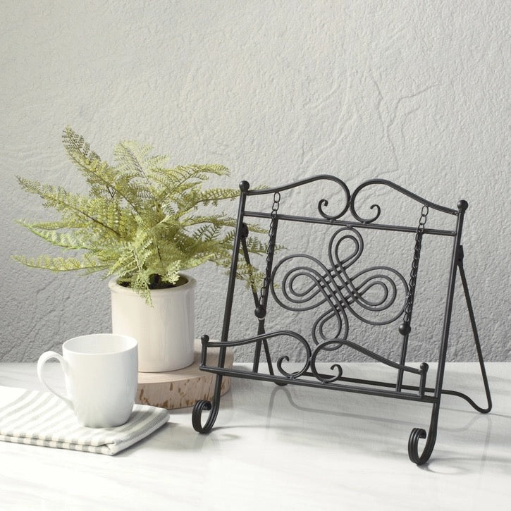 A Black metal decorative display stand with scrollwork and hanging rings, styled on a tabletop beside a potted fern, a white mug, and a striped napkin against a light gray textured wall.
