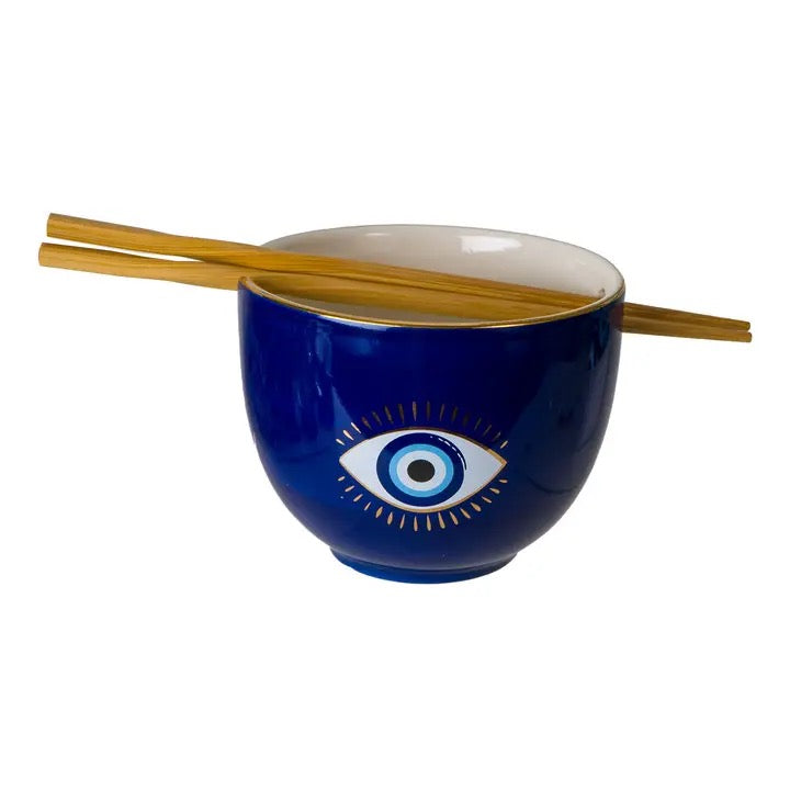 A dark blue ceramic bowl with a white interior and a blue-and-white evil eye design on the front, with two light brown wooden chopsticks resting across the top, shown on a plain white background.