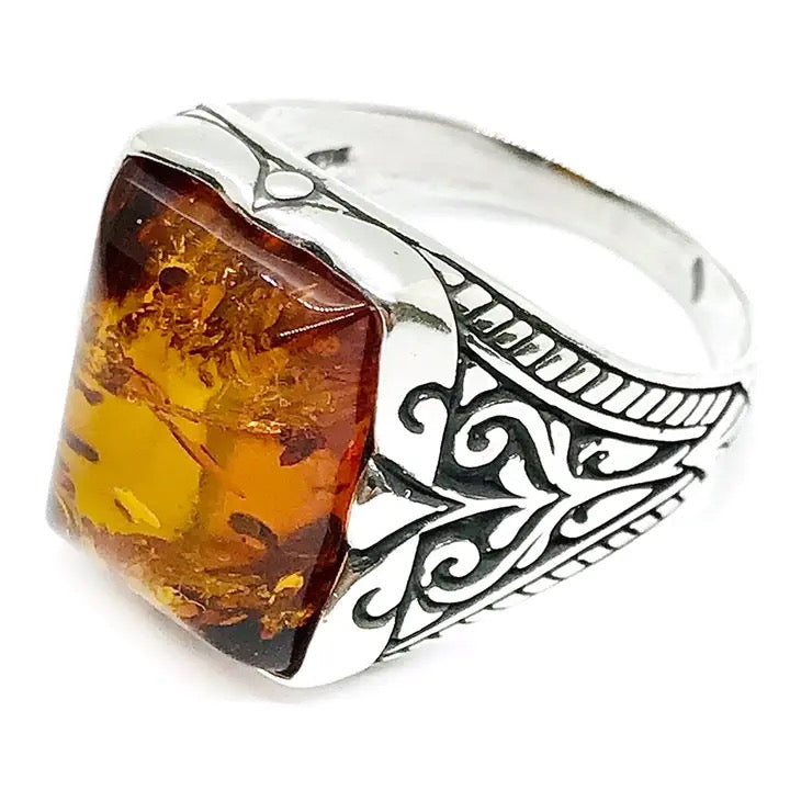 A sterling silver ring featuring a large, rectangular, bezel-set amber stone with intricate scrollwork on the band, shown against a plain white background.