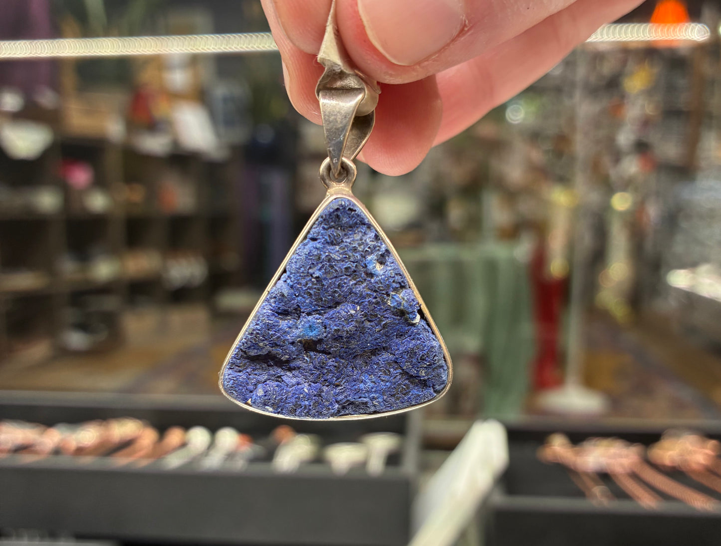 Triangular blue stone pendant held by a hand with a blurred jewelry store background