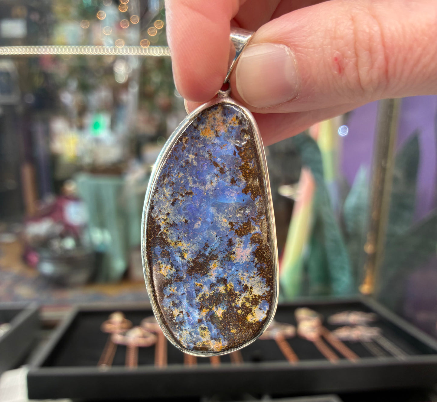 Hand holding a pendant with a blue and brown stone against a blurred jewelry store background