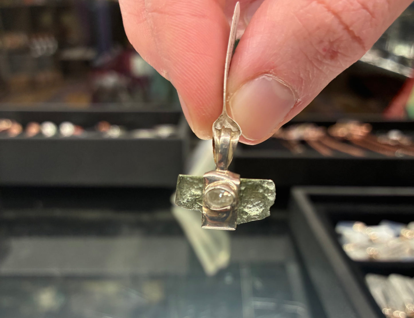 Close-up of a hand holding a silver pendant with a green stone against a blurred jewelry store background.