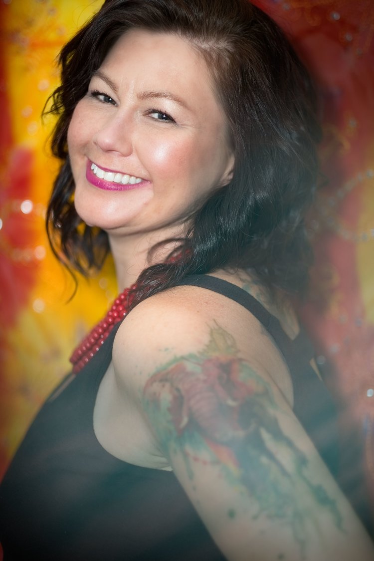 A smiling woman with dark hair and a colorful tattoo on her upper arm, wearing a black top and red beaded necklace against a vibrant yellow and red background.