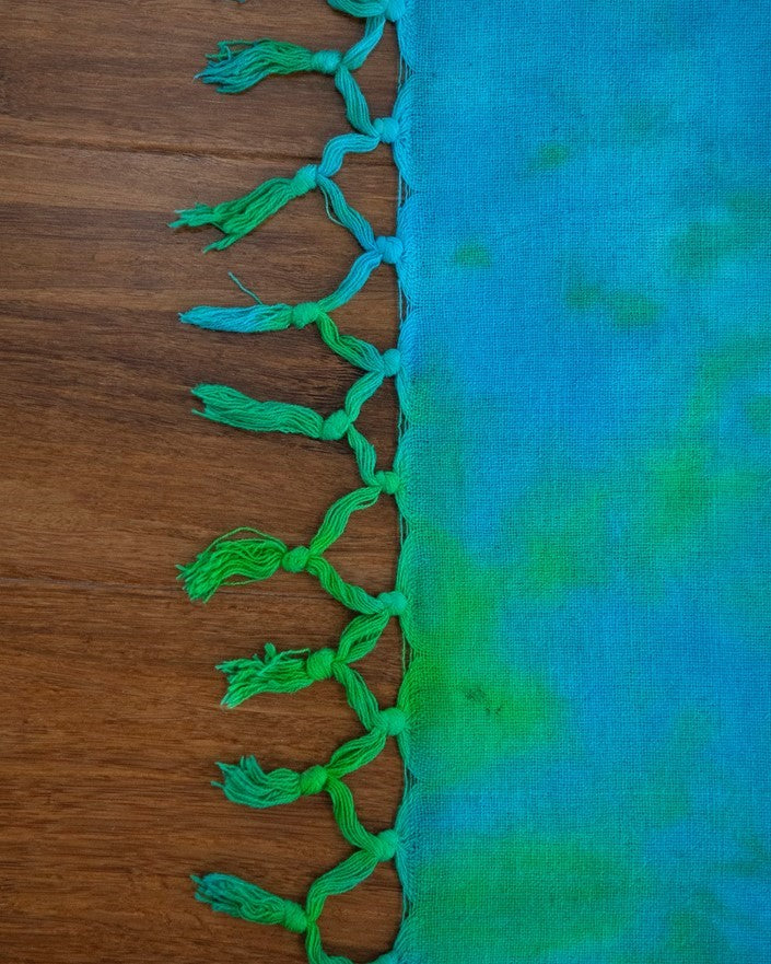A close-up of a fabric item, likely a scarf or throw, featuring a vibrant blue and green tie-dye pattern with a decorative border of green tassels, laid flat on a wooden surface.