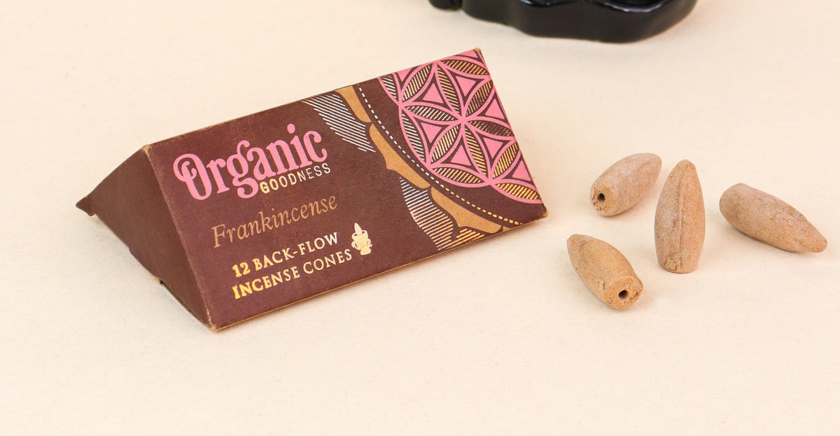 A triangular brown and pink box labeled 'Organic Goodness Frankincense' with four natural wood incense cones on a light beige surface.