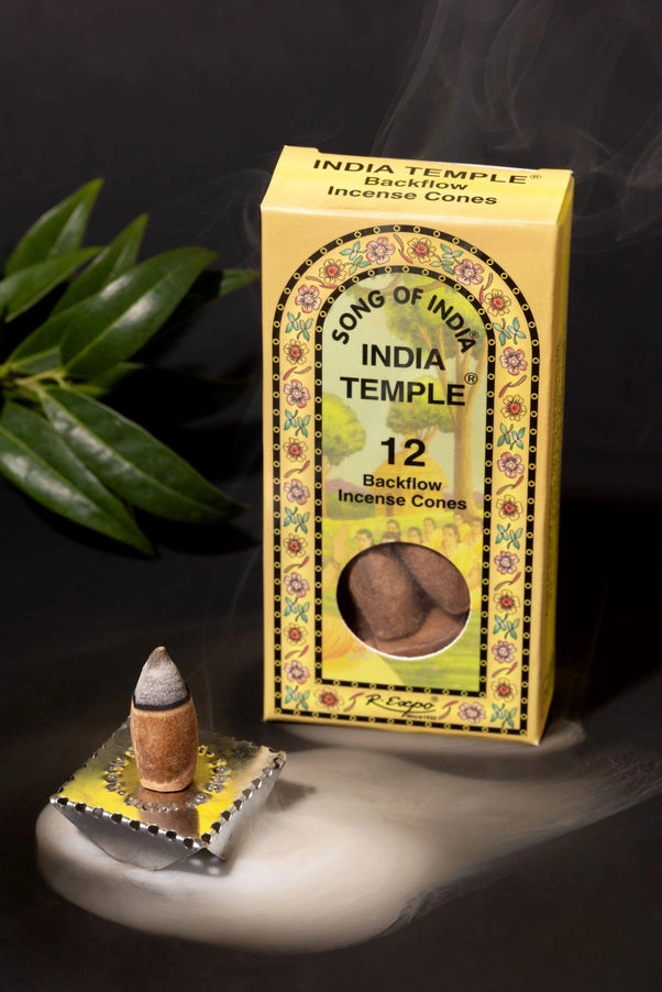A box of India Temple backflow incense cones stands on a white surface against a dark background, with a single incense cone resting on a small decorative plate in front and green leaves at the left edge.