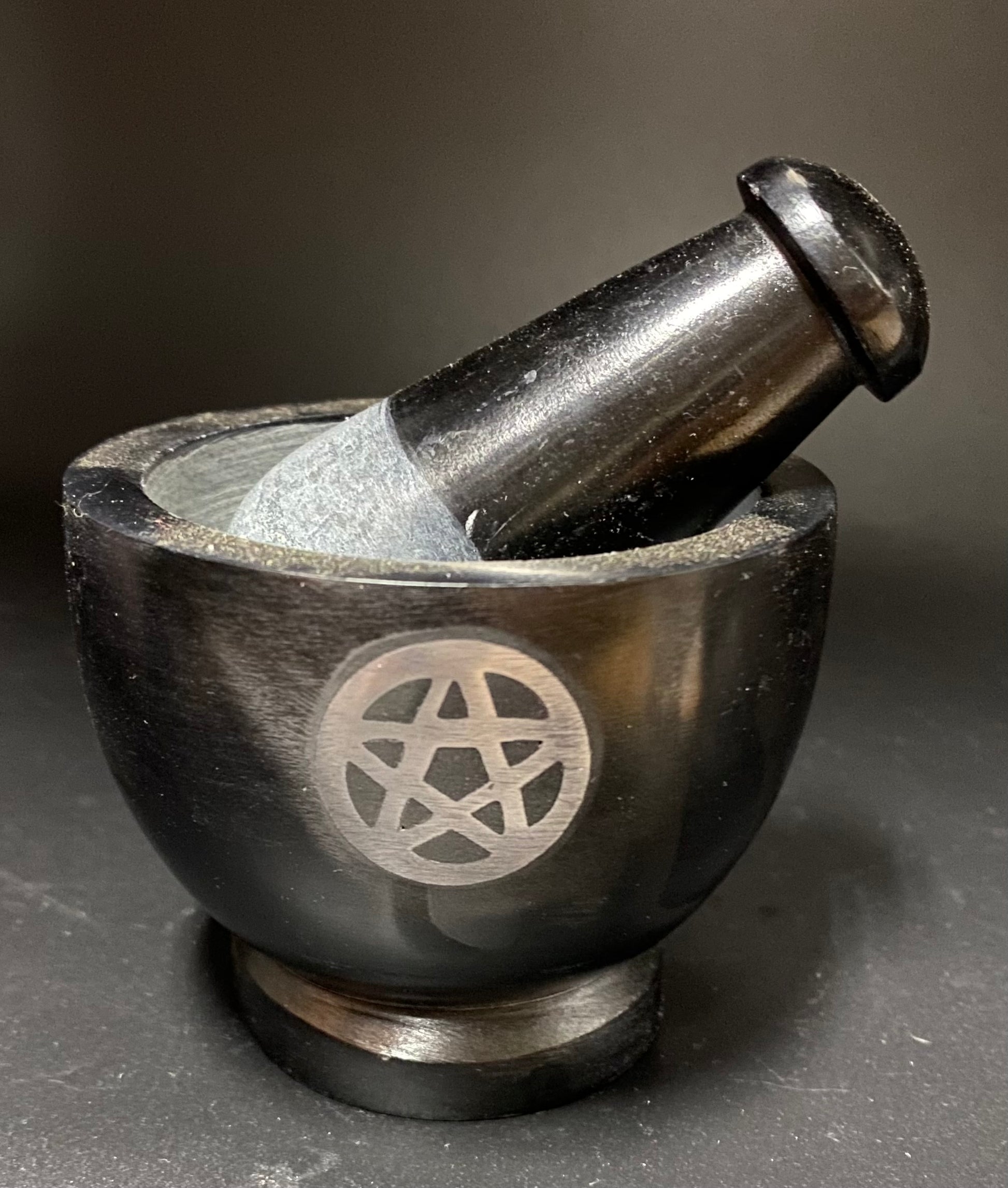 A black stone mortar and pestle set with a silver pentagram symbol engraved on the side, photographed on a dark gray background.