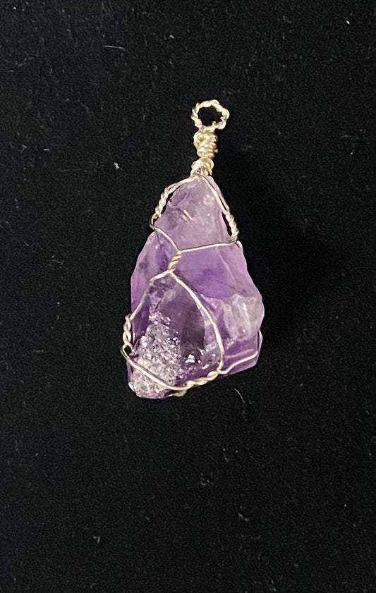 A raw purple amethyst crystal pendant wrapped in silver-toned wire, shown against a solid black background.