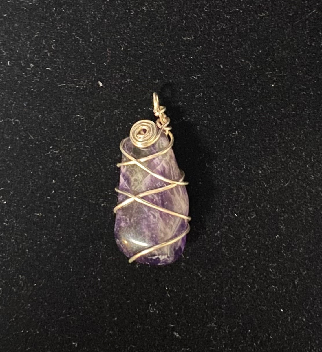 A raw purple amethyst stone pendant wrapped in silver-colored wire, displayed on a textured black background.