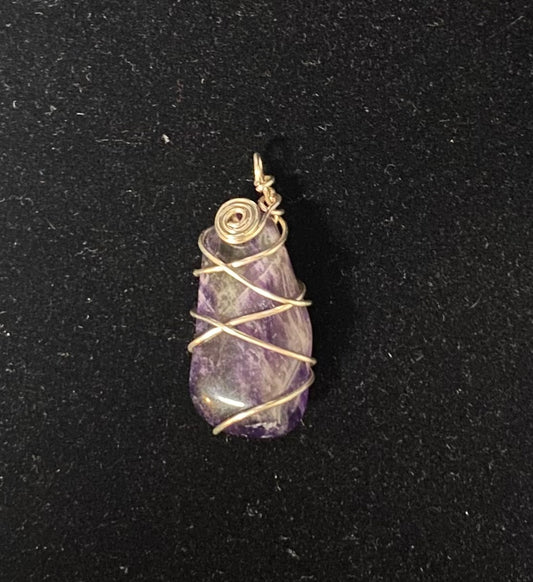 A raw purple amethyst stone pendant wrapped in silver-colored wire, displayed on a textured black background.