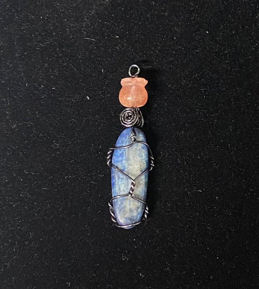 A handmade pendant featuring a blue kyanite wrapped in black copper wire, topped with a small strawberry quartz bead tulip wrap, set against a dark textured background.