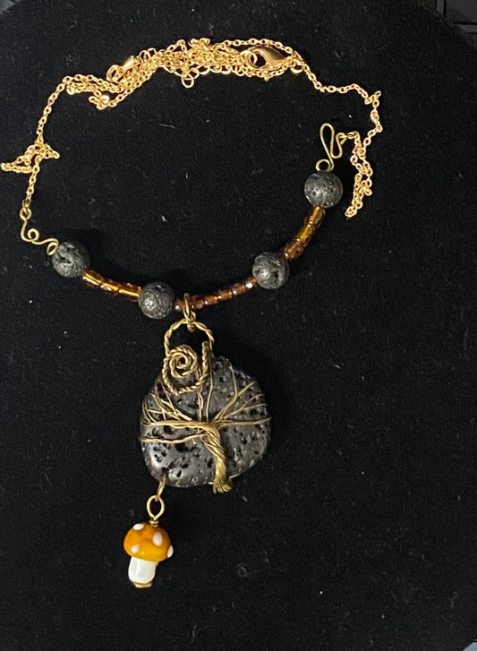 A handmade-style gold chain necklace with dark gray lava stone beads, a central lava stone wrapped in gold wire, and a small orange-and-white mushroom charm, displayed on a black background.