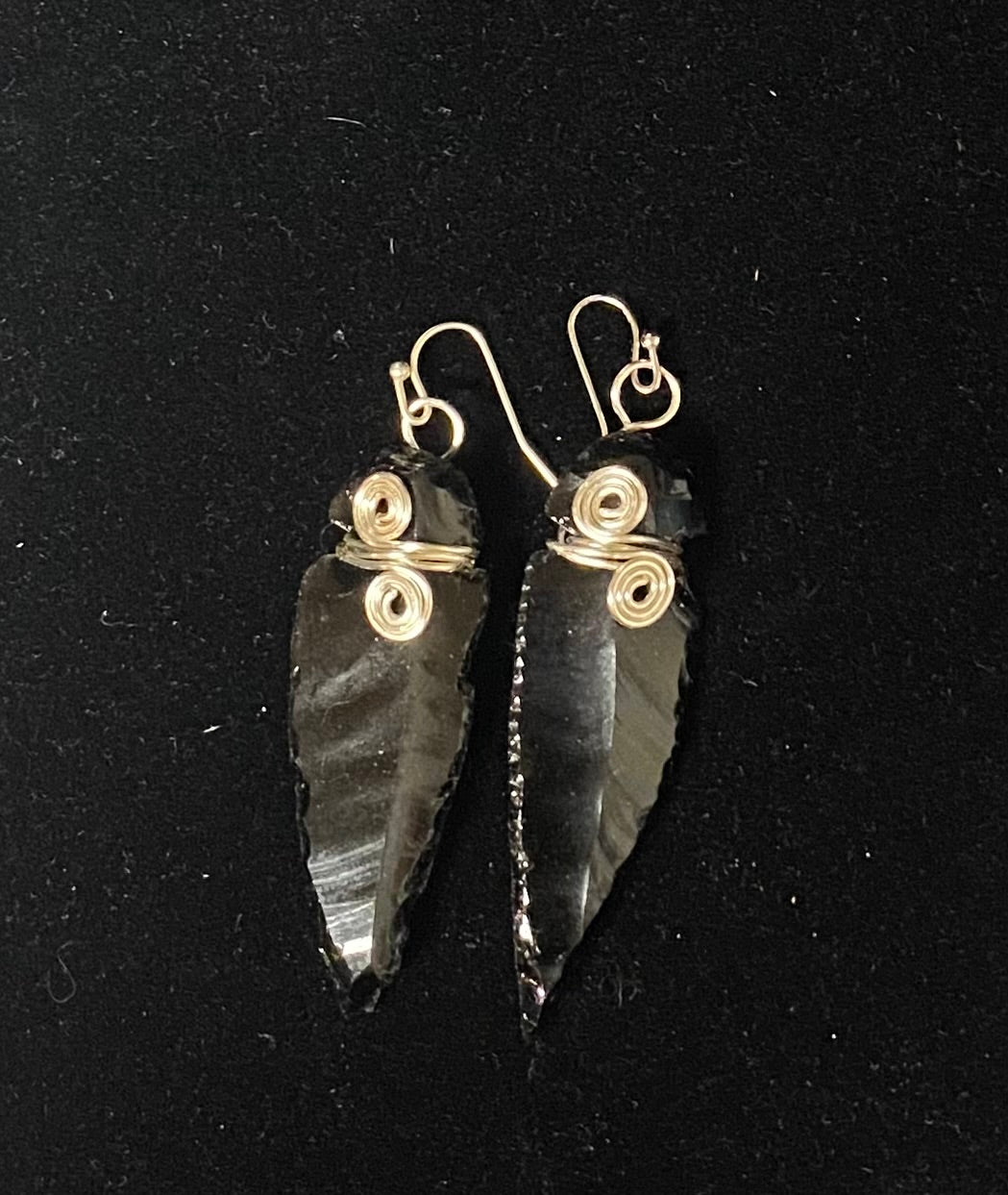 A pair of handmade dangle earrings featuring polished black obsidian stone arrowheads wrapped at the top with silver-toned wire coils and hooks, displayed on a black background.