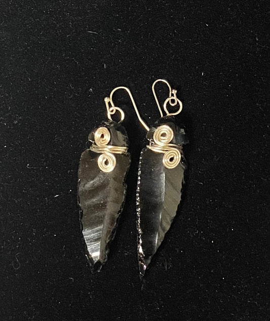 A pair of handmade dangle earrings featuring polished black obsidian stone arrowheads wrapped at the top with silver-toned wire coils and hooks, displayed on a black background.