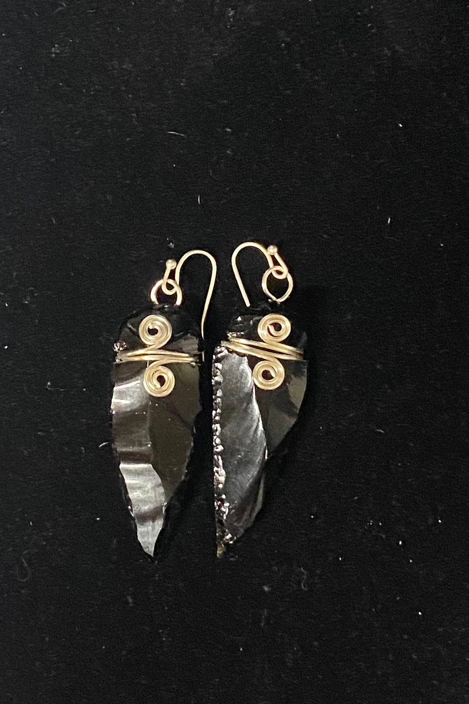 A pair of handmade dangle earrings featuring natural black obsidian arrowheads wrapped with silver wire spirals and hooks, displayed on a black background.