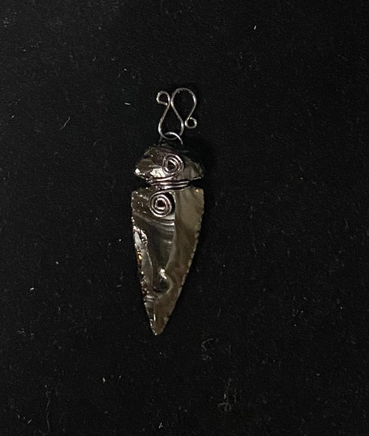 A handmade obsidian pendant arrowhead with textured swirls, attached to a simple wire loop, shown on a black background.