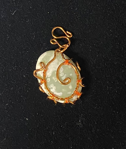 A handmade pendant featuring a light green jadeite stone wrapped in textured bronze and copper wire, set against a dark textured background.