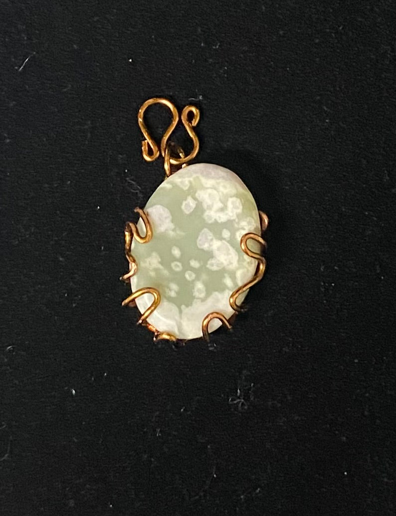 An oval pendant featuring a pale green and white marbled stone in a bronze wire-wrapped setting with a matching bronze bail, shown on a black background.