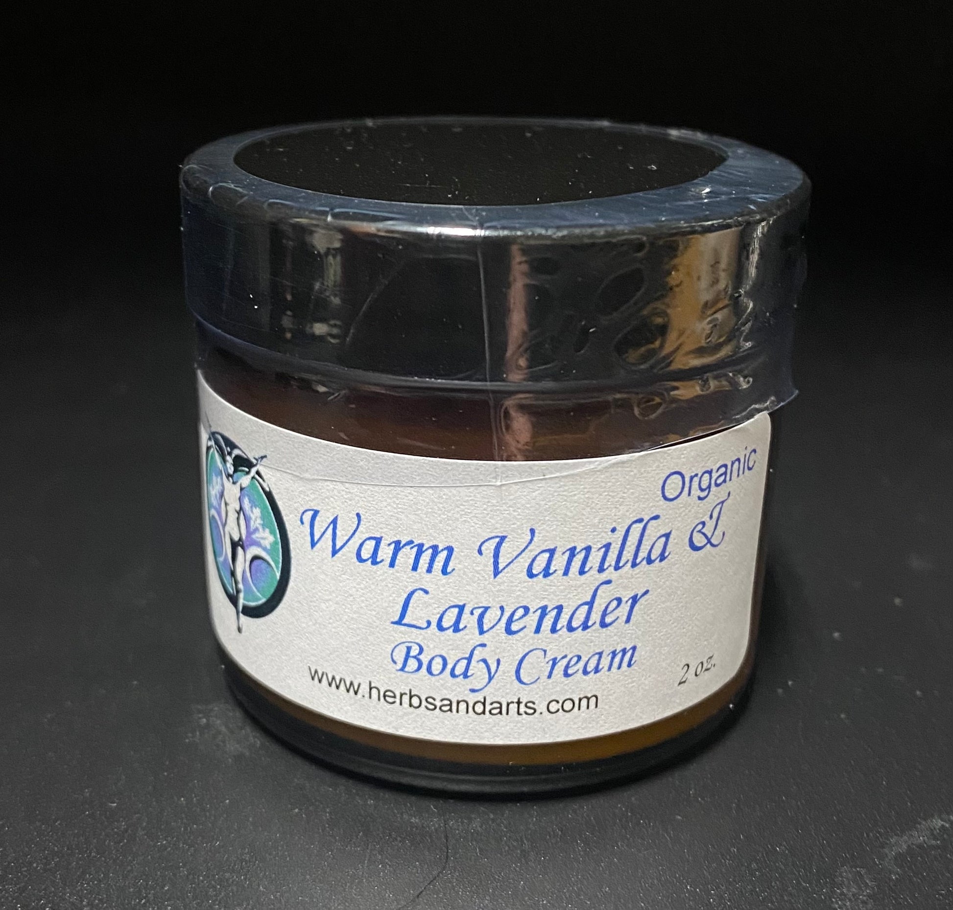 A small clear glass jar of body cream with a black lid and a white label reading 'Warm Vanilla & Lavender Body Cream' and 'Organic'.
