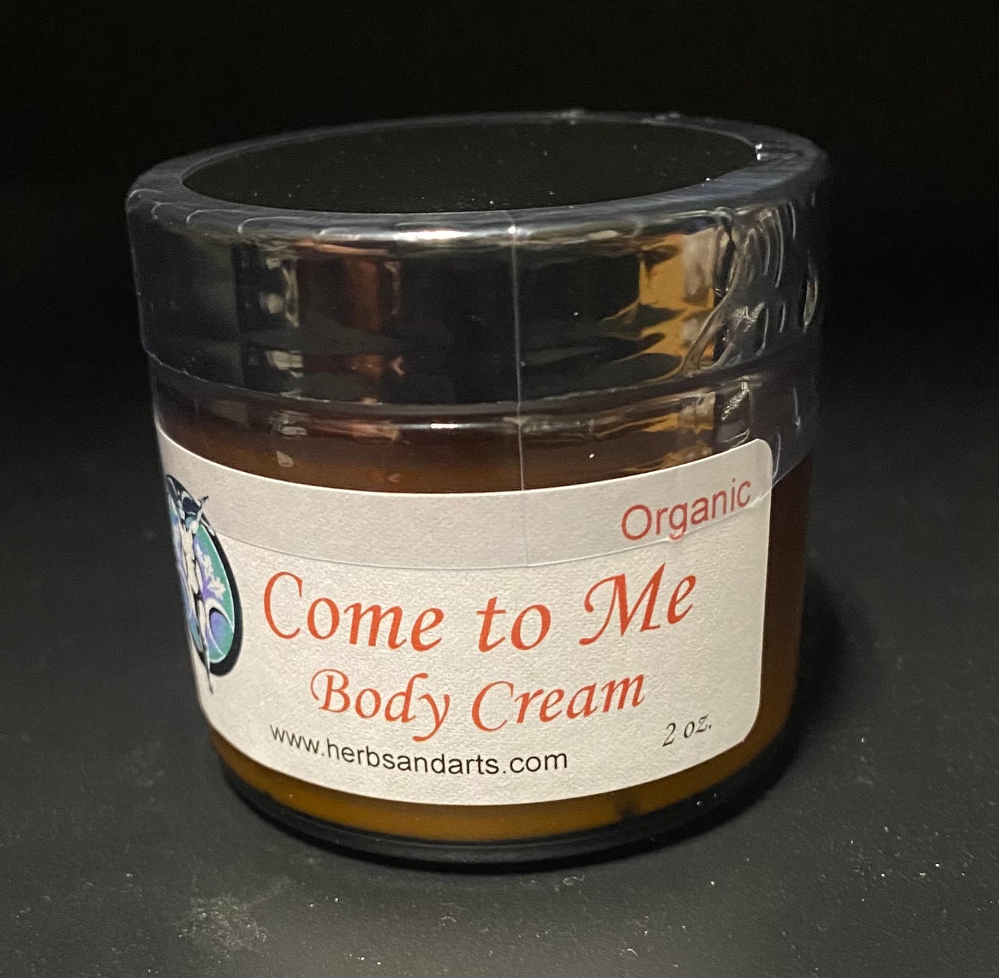A 2 oz jar of Herbs and Arts 'Come to Me' Organic Body Cream, featuring a white label with red text and a clear lid, set against a dark background.