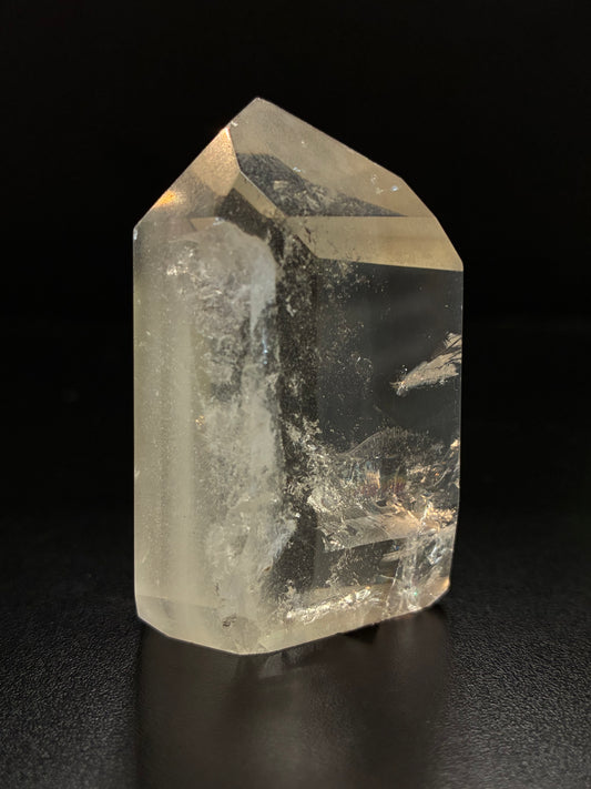 A natural clear quartz crystal point with internal inclusions, displayed on a dark reflective surface against a black background.