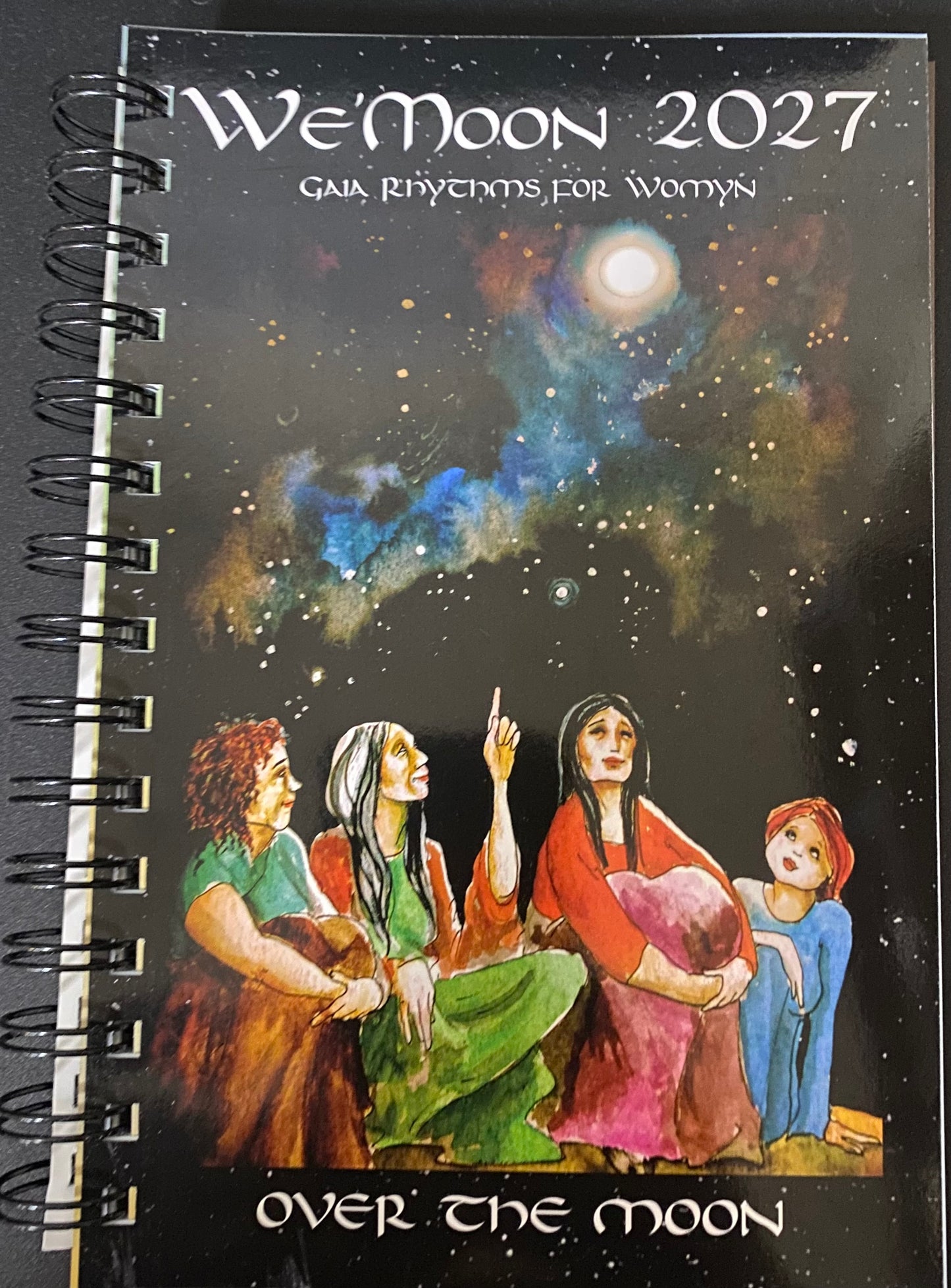 A spiral-bound notebook titled "We'Moon 2027" with the subtitle "Gaia Rhythms for Women" and the phrase "Over the Moon" on the cover. The cover art shows a dark starry sky with a glowing moon and an illustration of four women in colorful robes looking upward.
