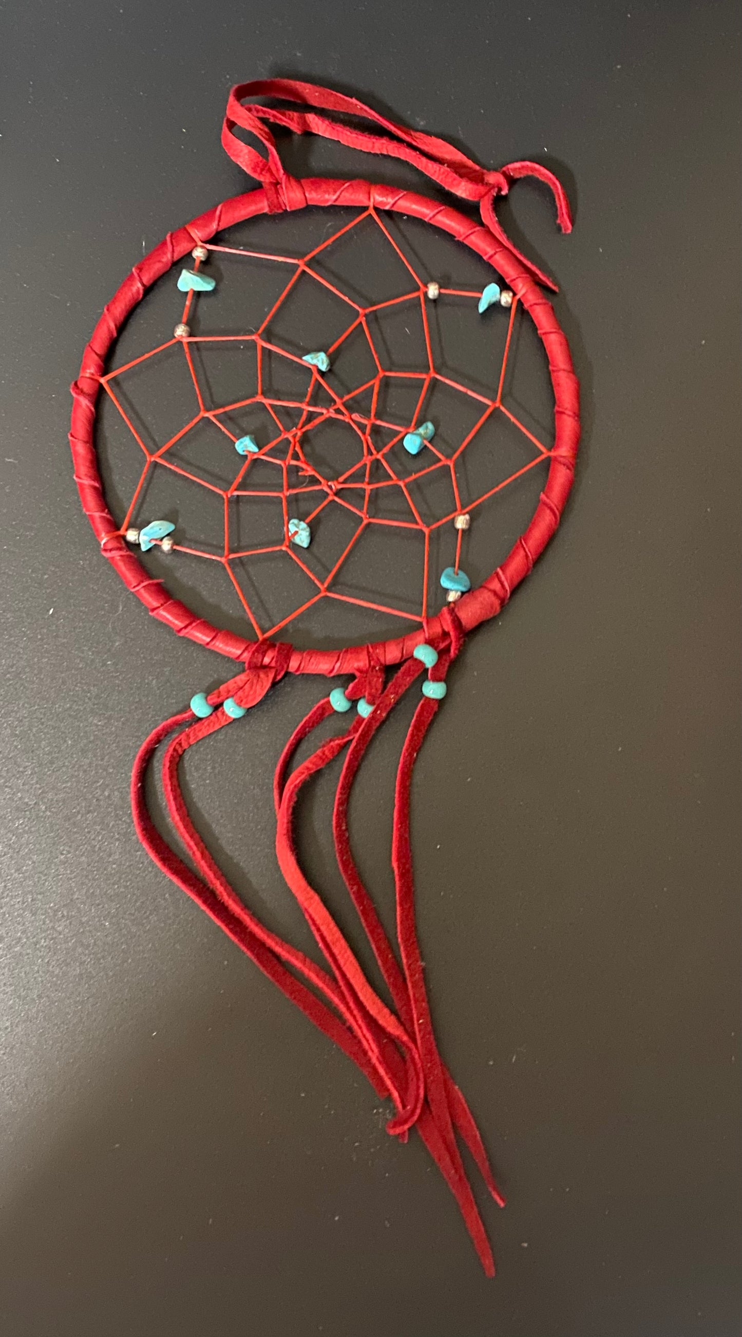 A handmade dreamcatcher with a red wrapped hoop, red webbing, and small turquoise-colored beads, shown on a dark gray background.