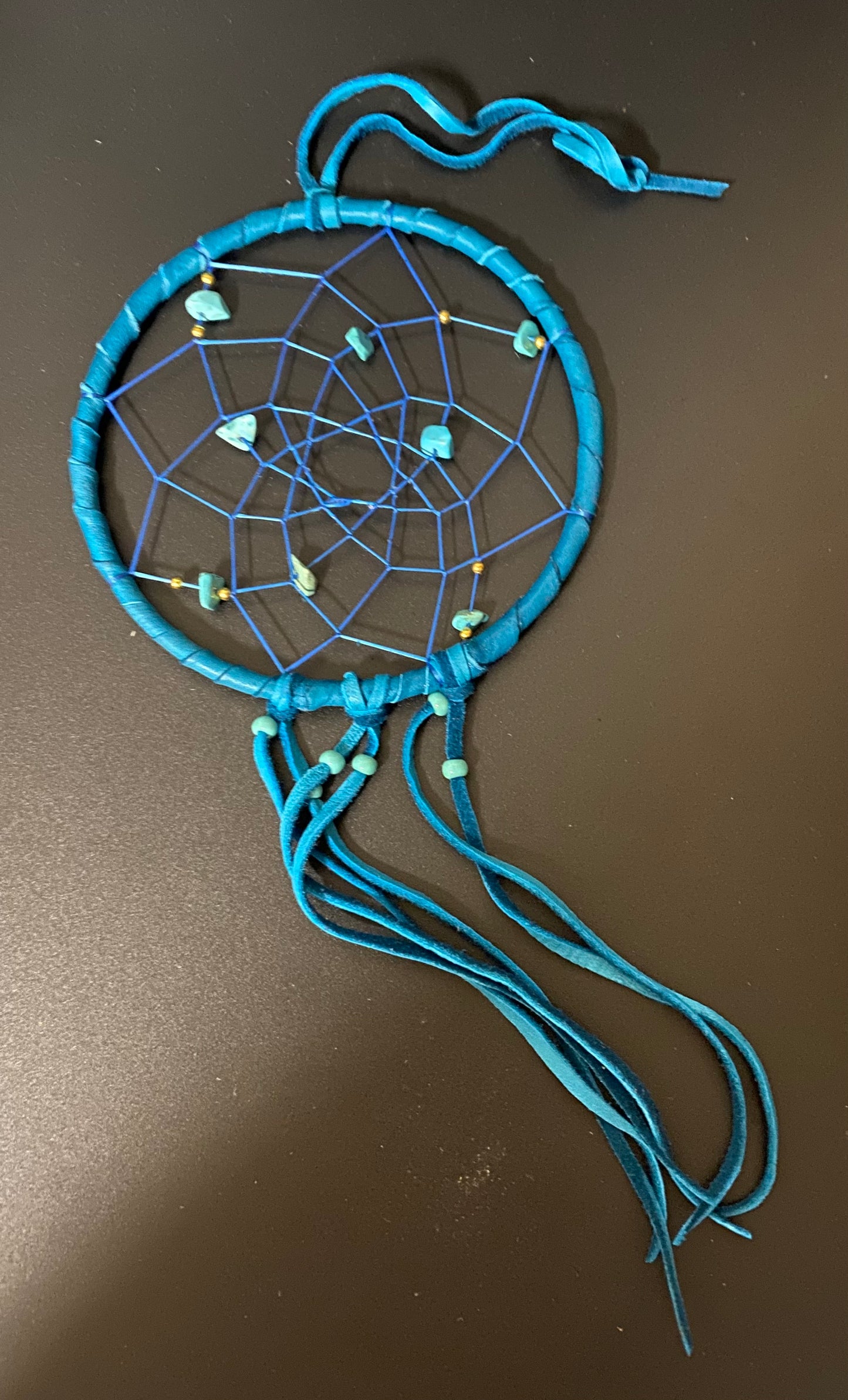 A handmade dreamcatcher with a bright blue wrapped hoop, blue webbing, and blue cords, decorated with small turquoise beads and natural stone charms, lying on a dark brown surface.