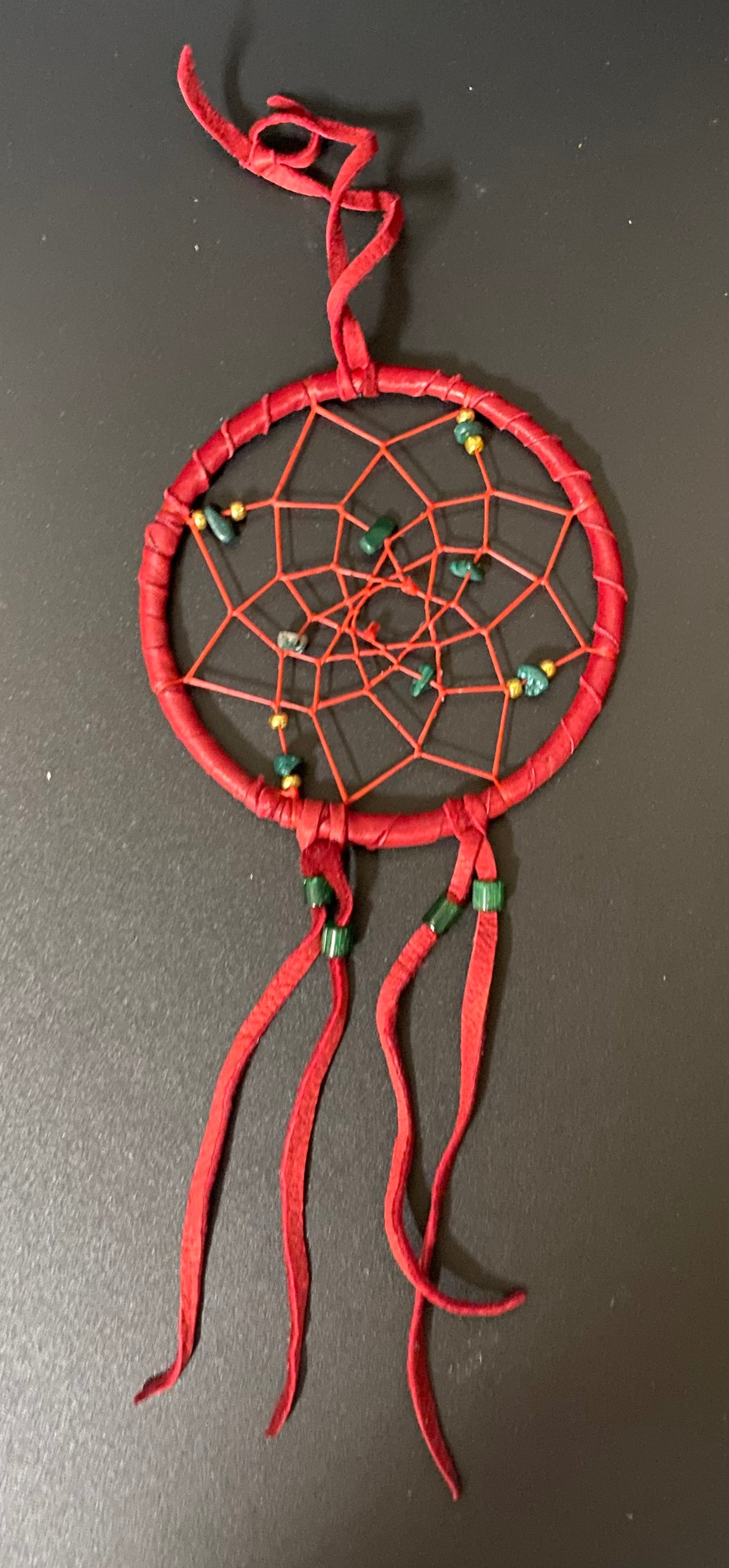 A handmade red dreamcatcher with a wrapped circular frame, RED webbing, and small green and yellow beads, hanging against a dark gray background.
