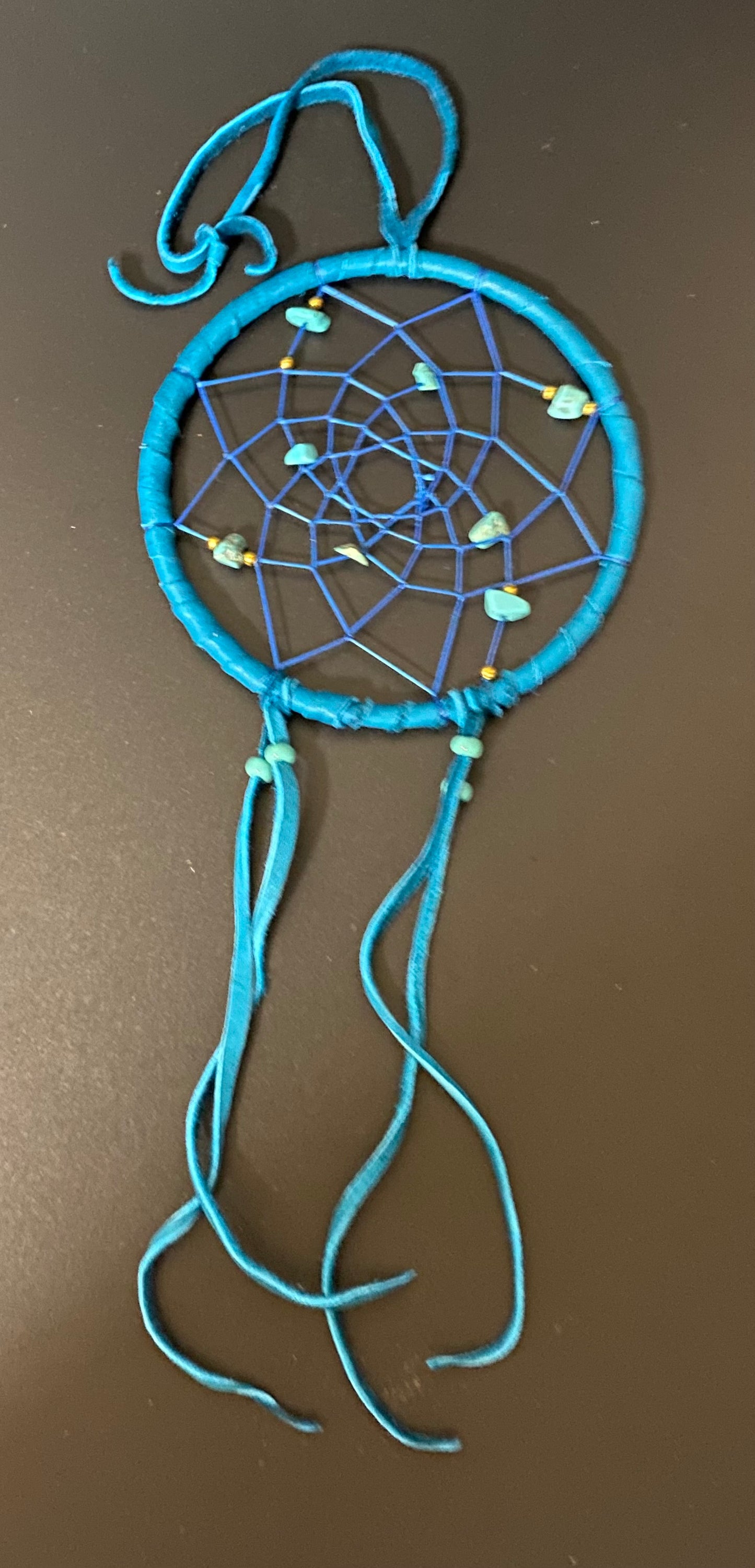A handmade dreamcatcher with a bright blue wrapped hoop, blue webbing, and long blue hanging tassels, decorated with small white and blue beads, shown against a dark brown background.