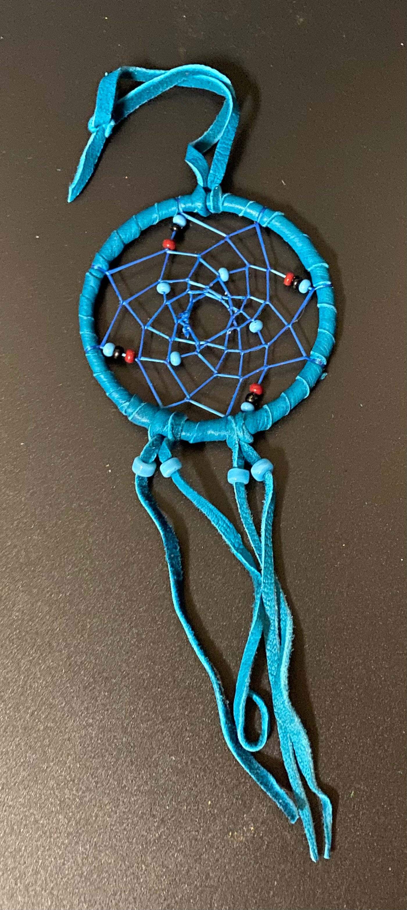 A handmade dreamcatcher made from bright turquoise blue suede ribbon, featuring a circular webbed center with small red and blue beads, photographed on a dark brown background.