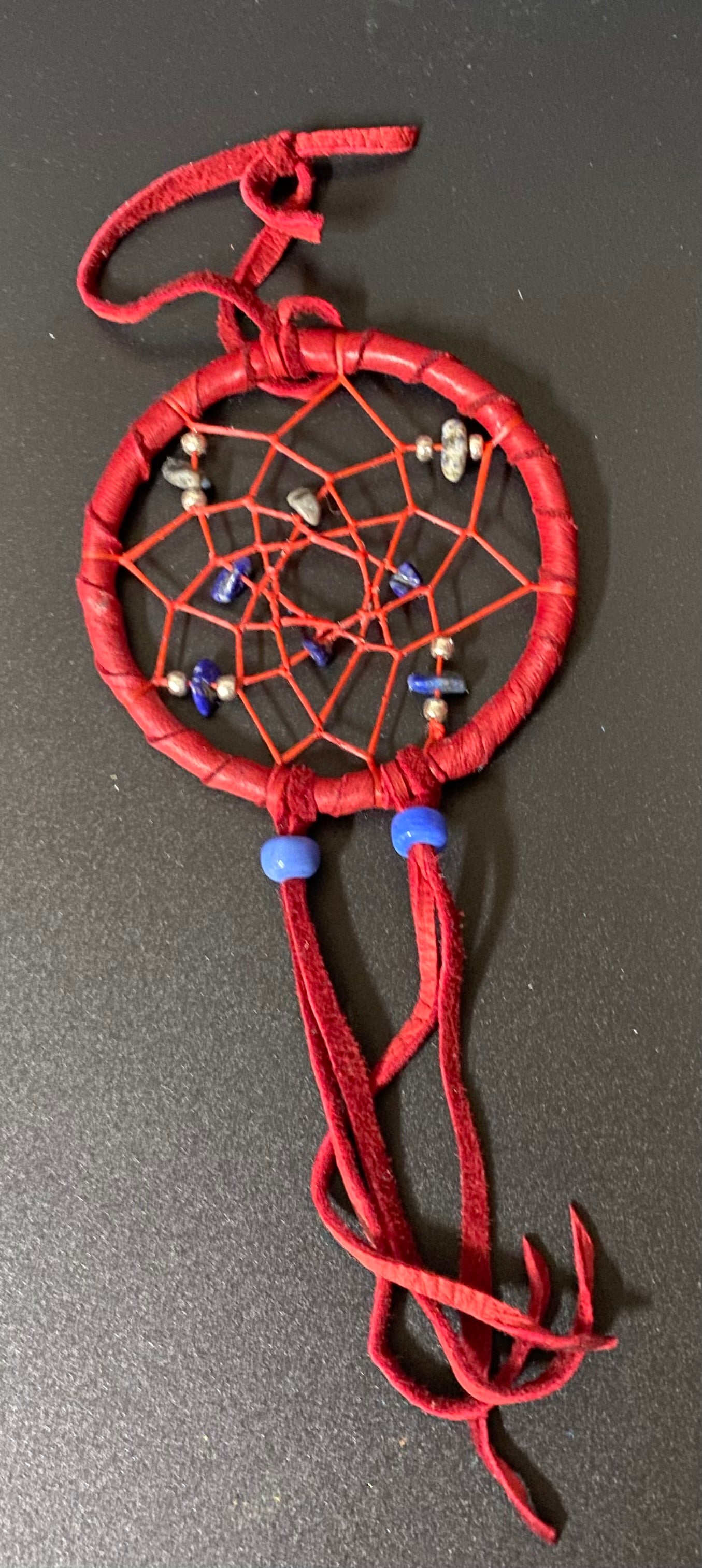 A red dreamcatcher ornament with a circular woven frame, red webbing, small decorative beads, and long red hanging tassels, photographed on a dark gray surface.