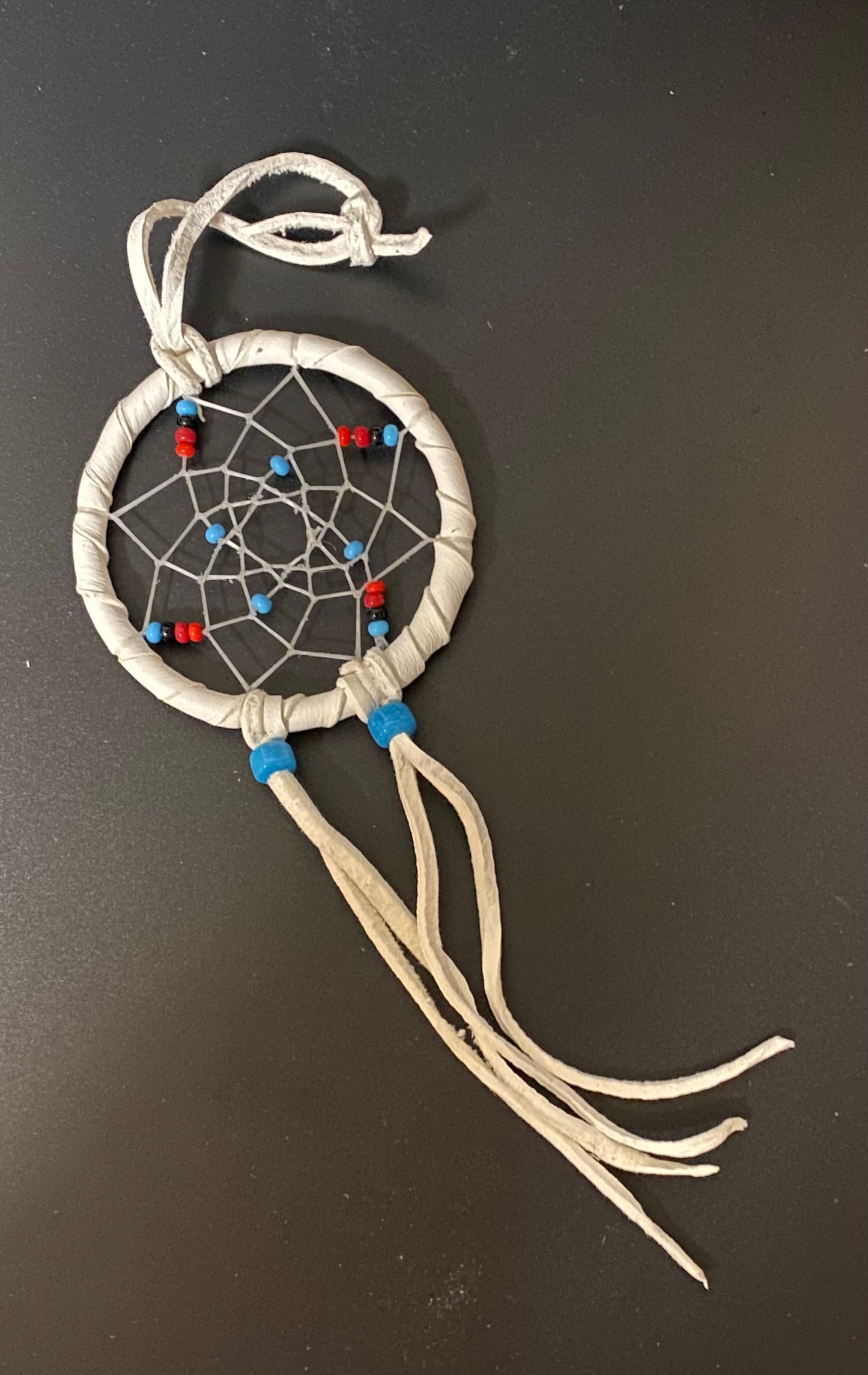 A small handmade dreamcatcher ornament with a white wrapped hoop, white webbing, and red and blue beads, photographed on a dark gray surface.