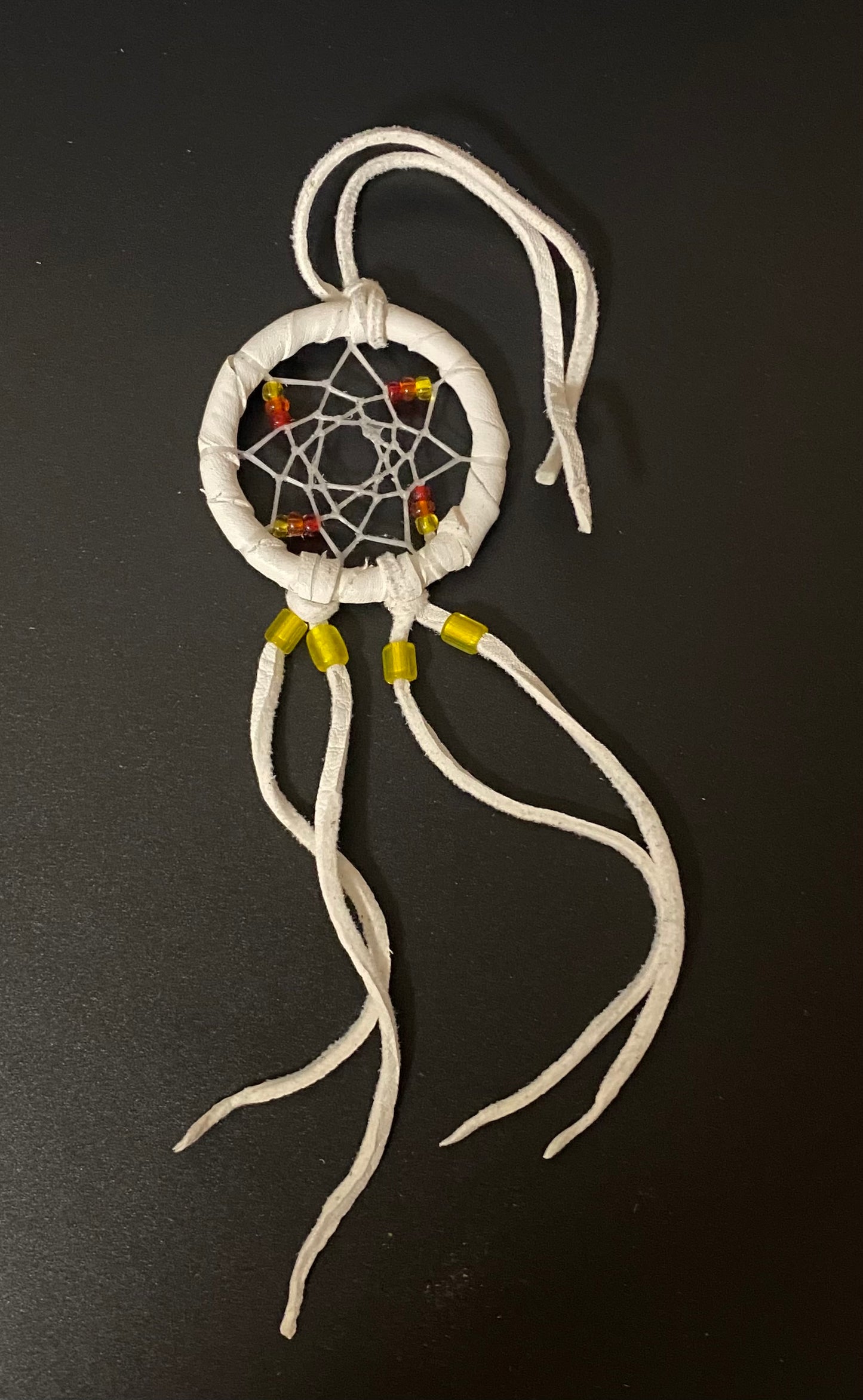 A small white dreamcatcher with white wrapped cord, white webbing, and small yellow and red bead accents, photographed on a dark background.