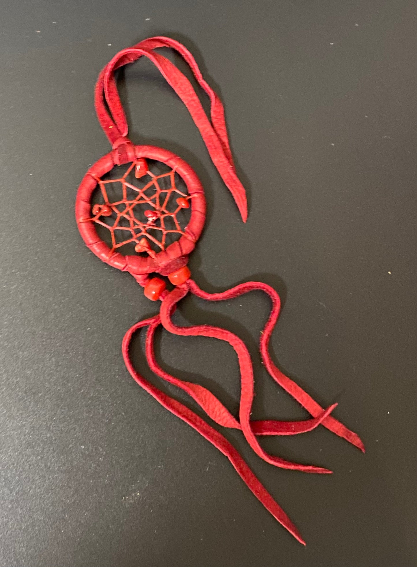 A small red dreamcatcher ornament made from wrapped cord or leather, with a web pattern in the center, small beads, and long hanging ties, photographed on a dark gray surface.