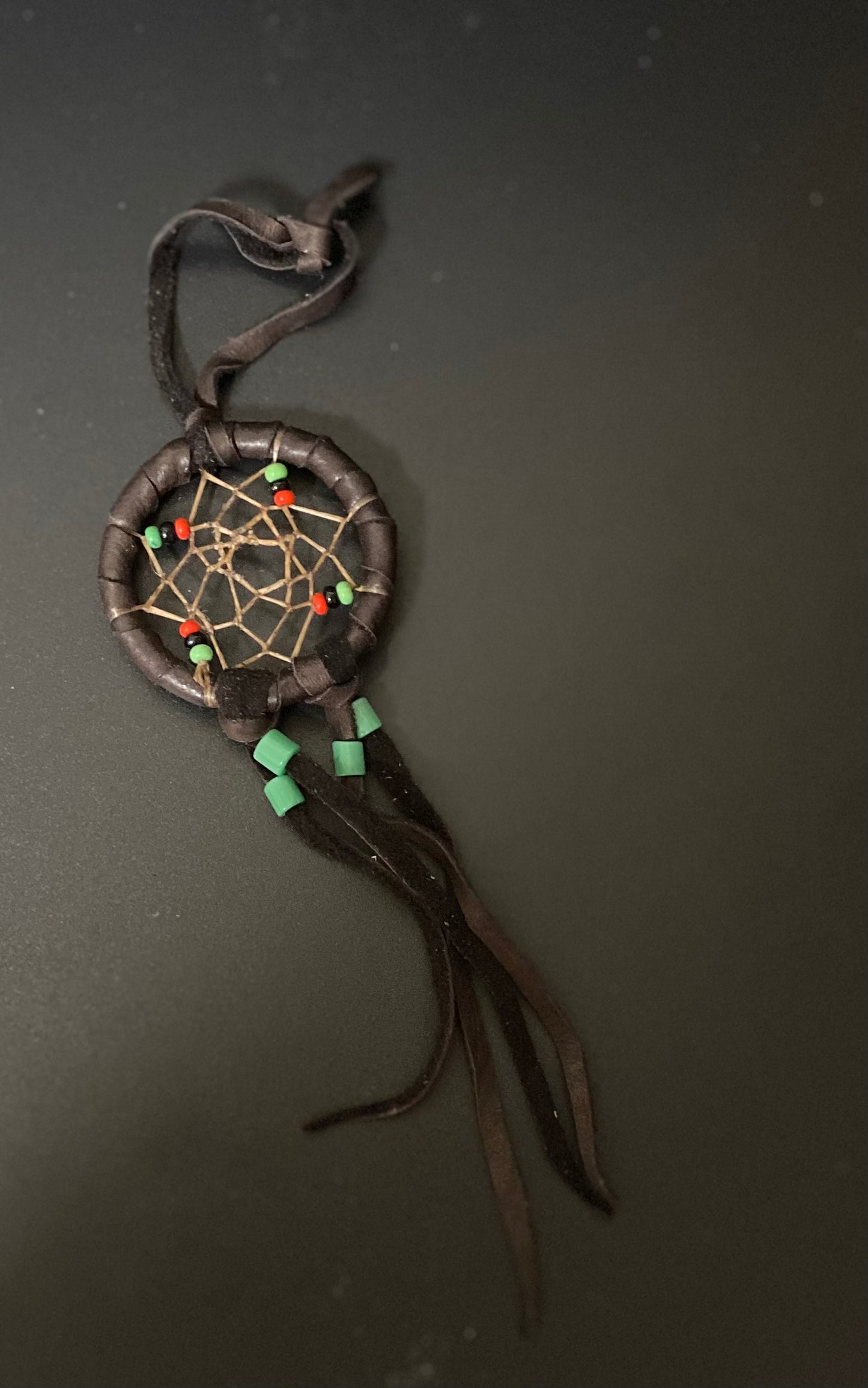 A small handmade dreamcatcher with dark brown leather wrapped body, tan webbing, small red and green beads, and dangling dark brown leather strands, photographed on a dark gray background.