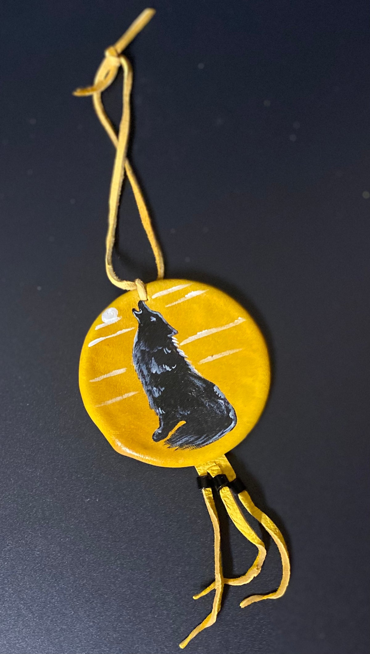 A handmade circular yellow shield  featuring a black painted wolf silhouette and white painted lines, hanging from yellow cords against a dark background.