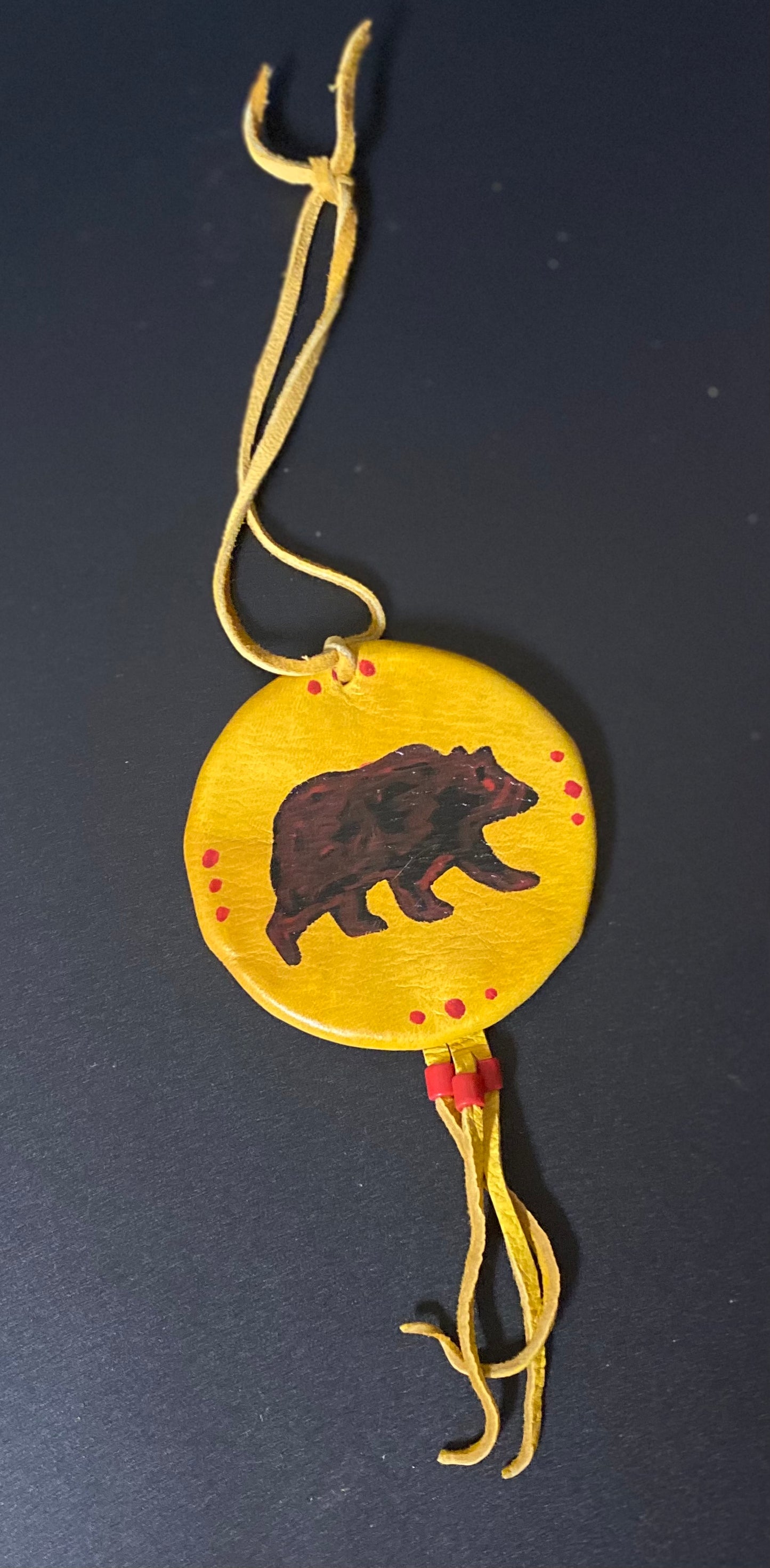 A circular yellow shield featuring a hand-painted brown bear silhouette in the center, surrounded by small red dots, hanging from a yellow cord against a dark background.