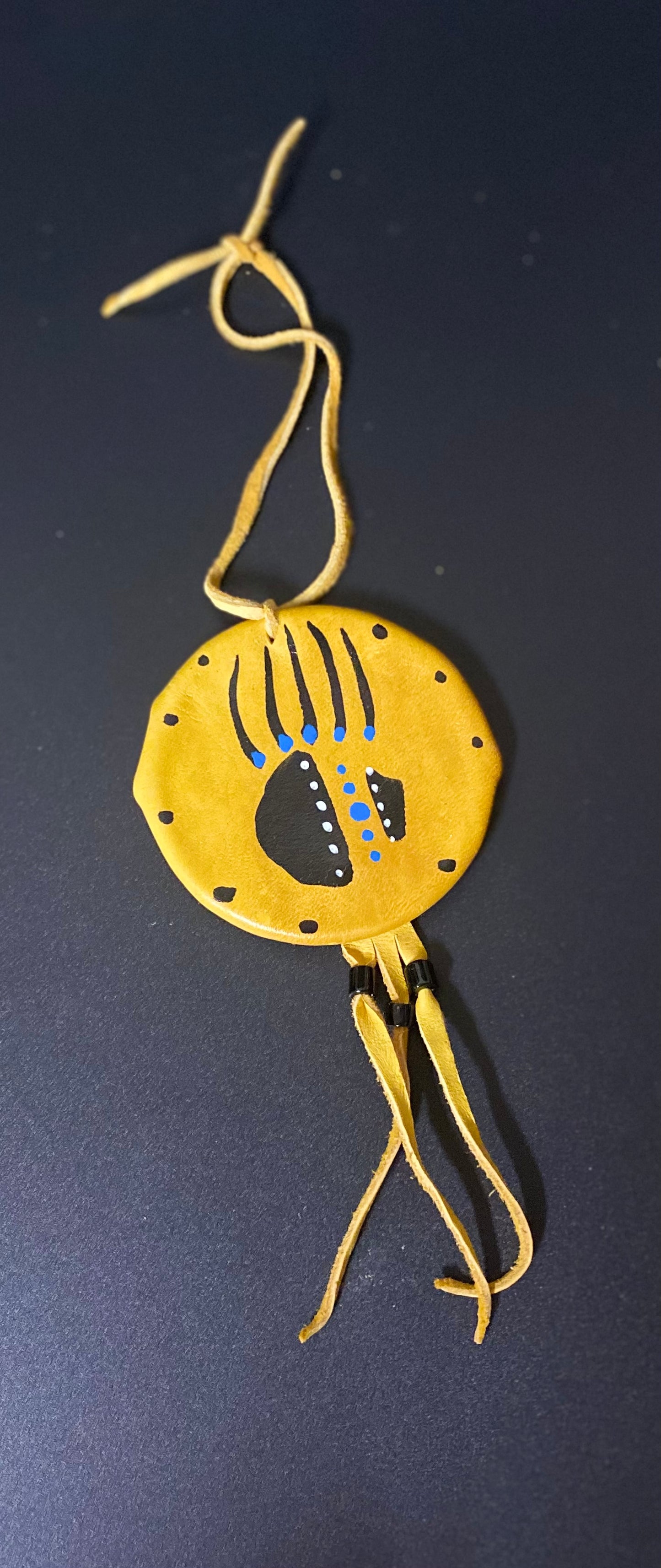 A yellow circular leather shield with a hanging cord, featuring a black and blue handprint design on the front, photographed against a dark background.