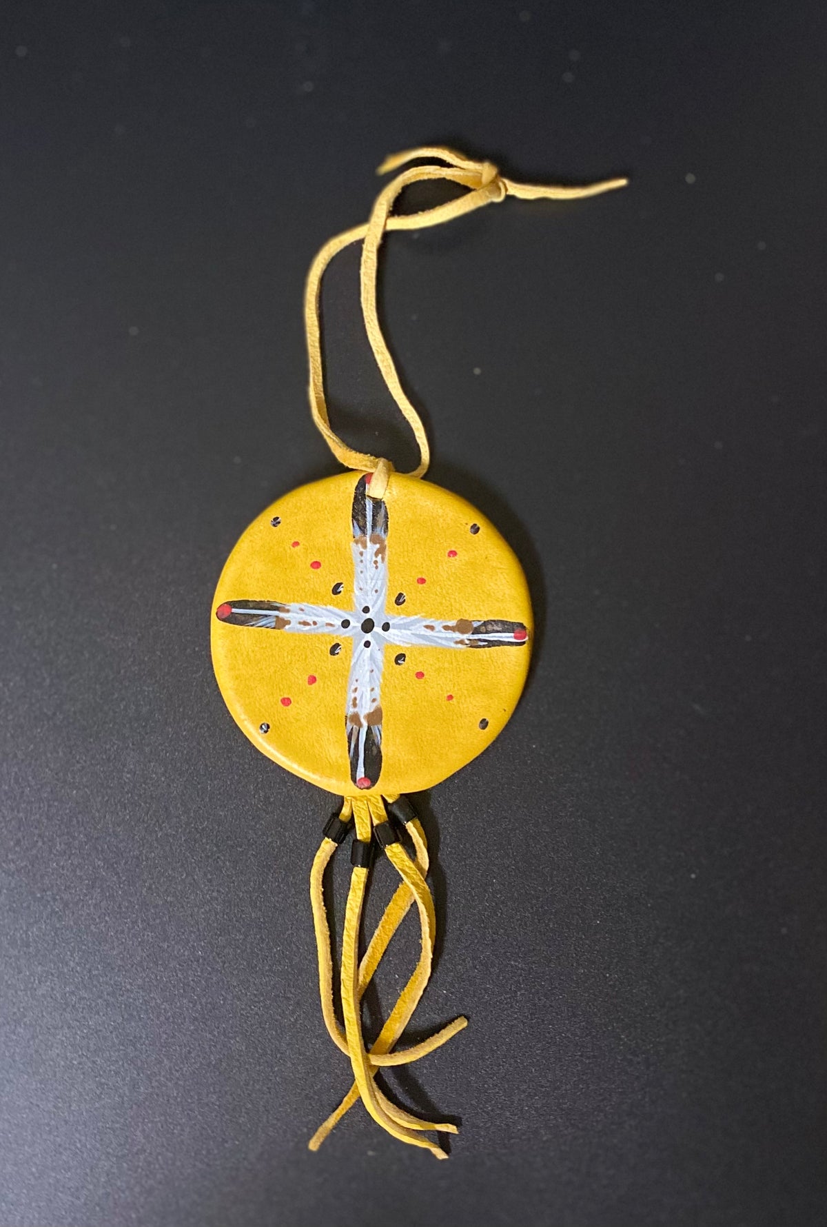 A circular yellow leather shield with a hand-painted white cross, red dots, and black accents, hanging from yellow cords against a dark gray background.