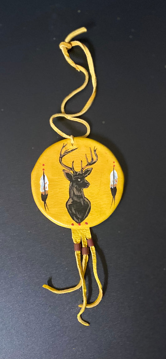 A circular yellow handcrafted shield featuring a black painted deer head and two feather details, suspended by a yellow cord on a dark background.