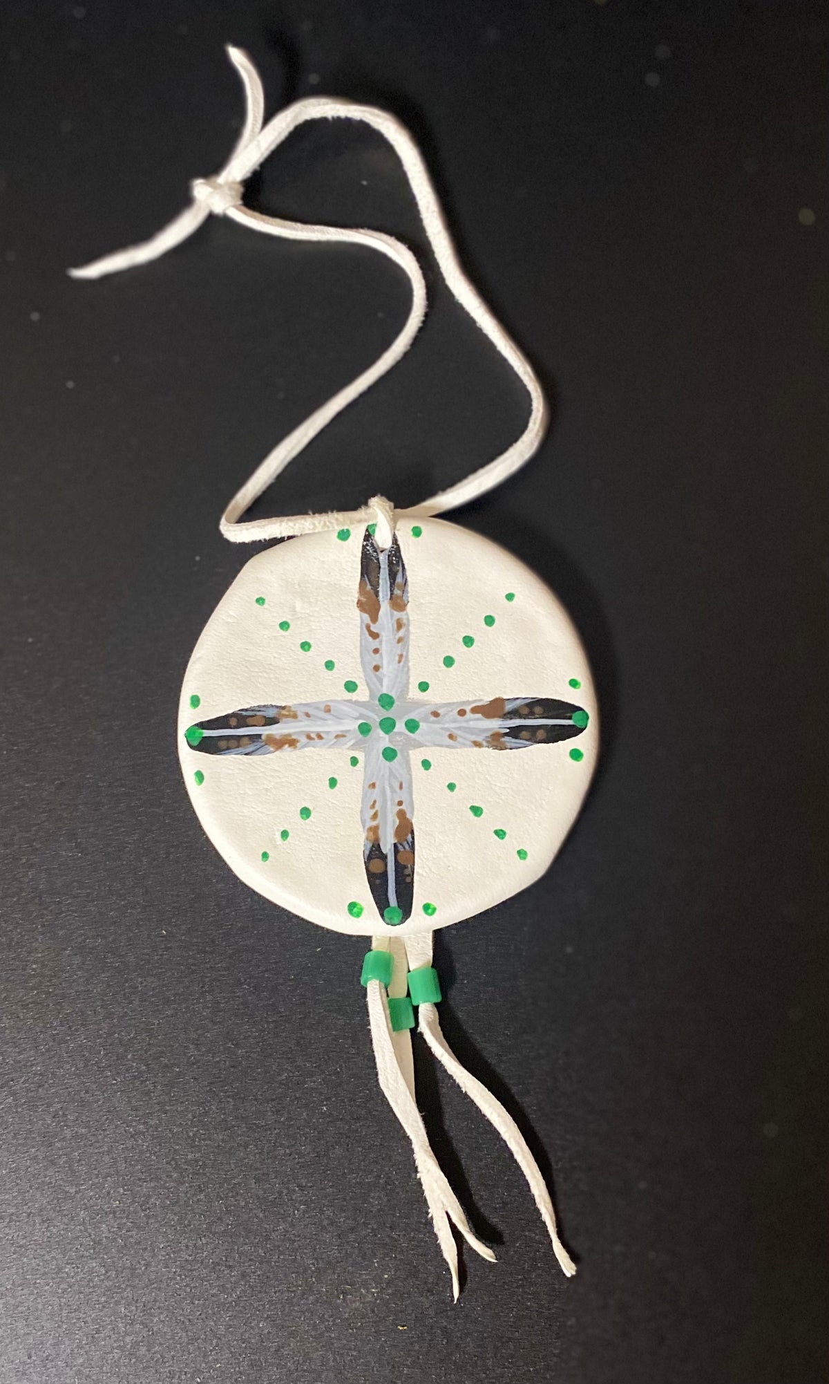 A circular white shield with a painted cross design in gray, black, and brown, accented with green dots and beads, hanging from a white cord against a dark background.